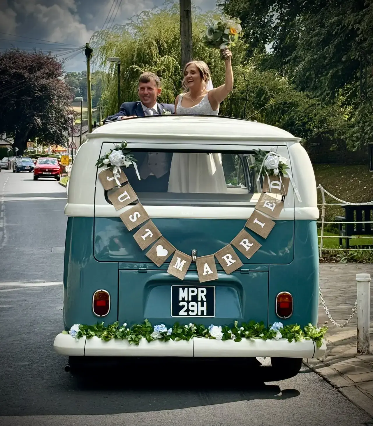 Jenny and her husband waving through the open roof of the blue and cream VW camper wedding car with a Just Married banner