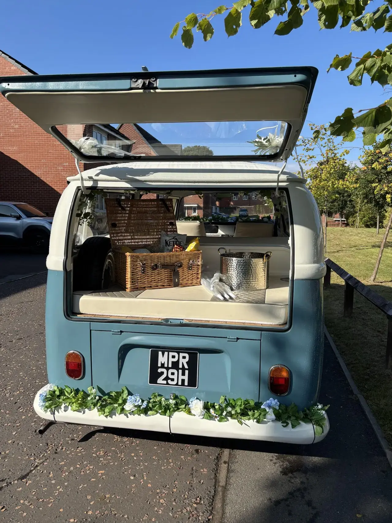 Open tailgate of the VW camper wedding car with a wicker picnic hamper, ice bucket and flower garland