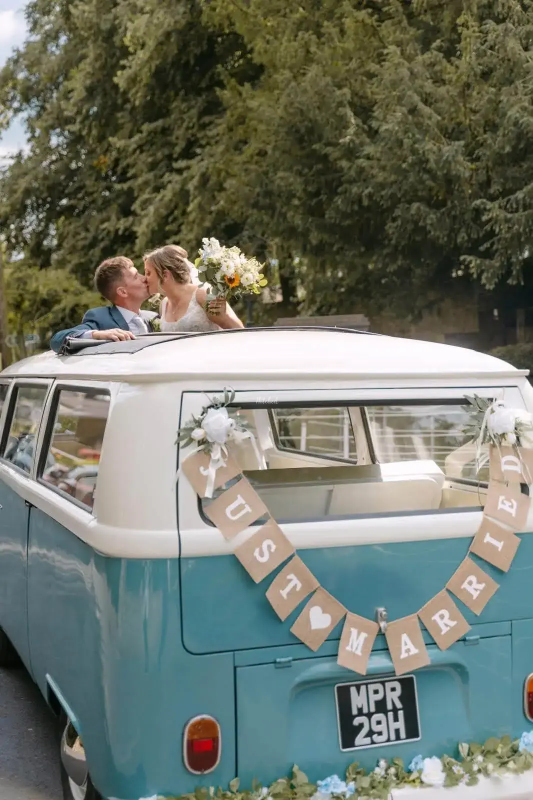 Bride and groom kissing through the open roof of the blue and cream VW camper wedding car with a Just Married banner